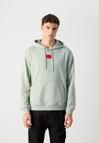 Joven con una sudadera verde clara que lleva una placa del logo rojo "HUGO" y pantalones negros, de pie contra un fondo sencillo.