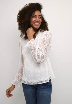 White blouse with intricate lace pattern, long sleeves with ruffled cuffs, and a relaxed fit, paired with denim jeans.