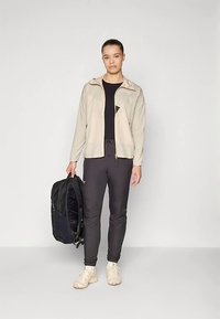 Gaiši bēša jaka ar rāvējslēdzēju, melns tops, melnas bikses un krēmkrāsas sporta apavi. Melna mugursoma turēta vienā rokā. Vienkāršs un funkcionāls dizains.