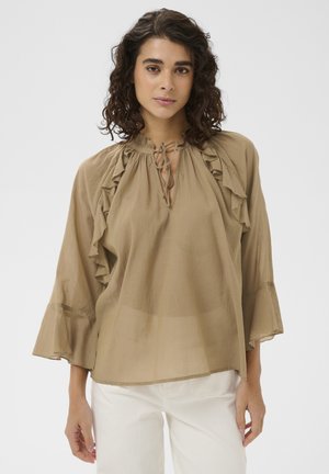 Žena s kučeravými vlasmi, oblečená v voľnej svetlohnedej blúzke s volánikmi a viazaním na krku, v kombinácii s bielymi nohavicami, stojaca pred jednoliatym pozadím.