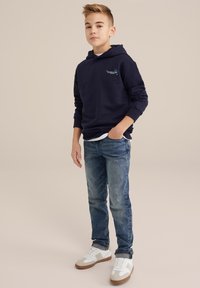 Felpa con cappuccio blu navy con tasca frontale, abbinata a jeans azzurri e sneakers bianche con dettagli grigi. Design semplice, vestibilità casual.