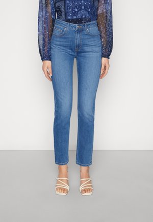 Kvinde iført lyseblå jeans med lige ben, en blå mønstret, gennemskinnelig langærmet top og hvide sandaler med remme på en grå gulv.