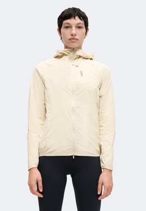 Person trägt eine hellbeige Kapuzenjacke, die zugezippt ist, und steht mit entspannten Armen vor einem einfarbigen hellgrauen Hintergrund.
