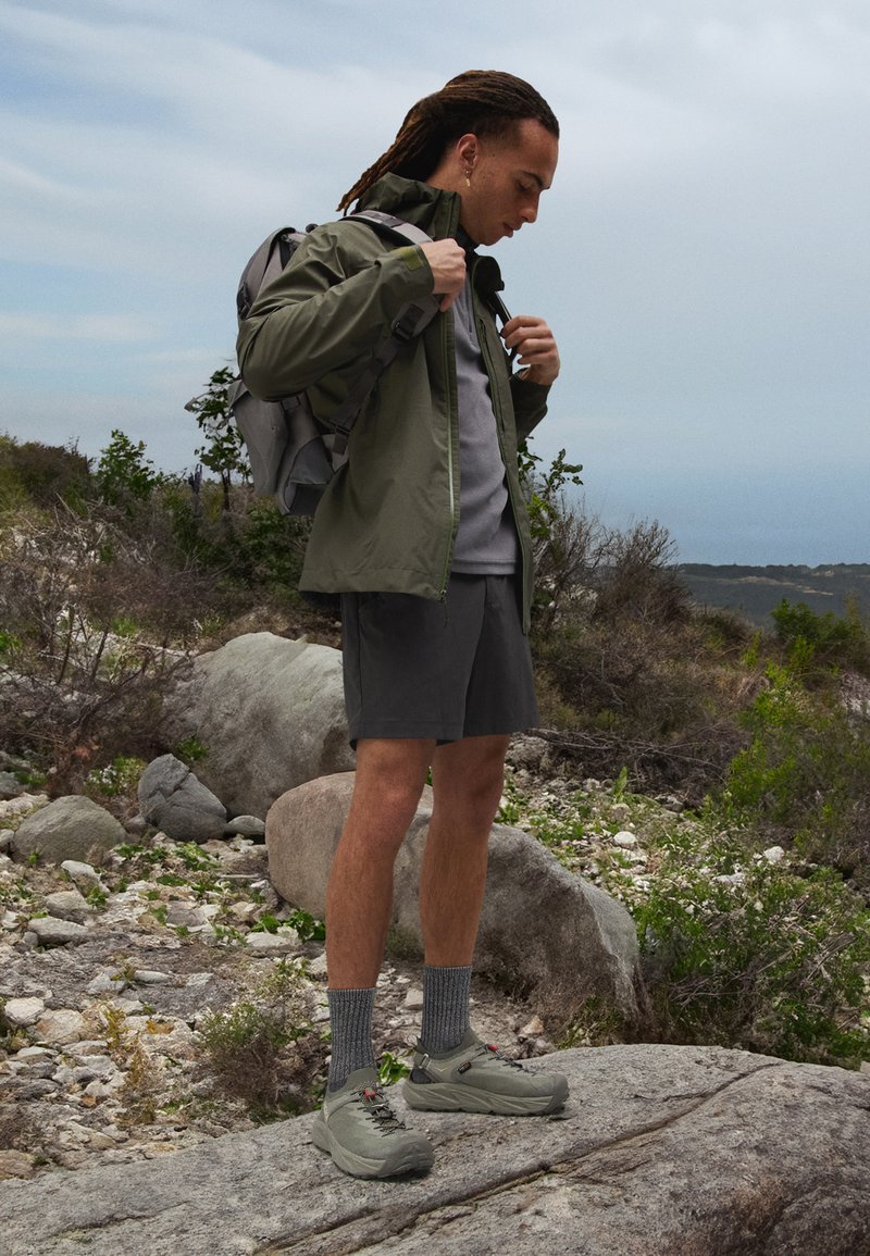 Zielona wodoodporna kurtka, szary t-shirt, ciemne spodenki i szare buty trekkingowe z teksturowanymi skarpetami. Noszona plecak, skaliste otoczenie na świeżym powietrzu.