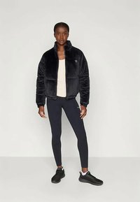 Juoda dirbtinė kailio striukė su aukštu apykakle, turi užtrauktuką priekyje ir logotipo detalę. Sujungta su juodomis leggingomis ir juodais sportiniais batais.