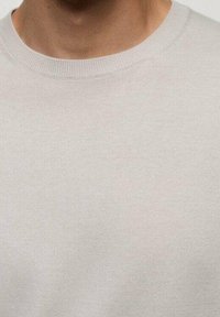 Pull gris clair avec une texture lisse, col rond côtelé, sans motifs ni accents visibles. Gros plan sur le détail du tissu sans ornements.