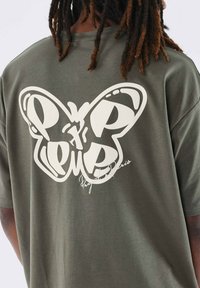 Camiseta verde oliva con un gran gráfico de mariposa blanca en la espalda y detalle de firma en cursiva. Tejido suave y ligero.