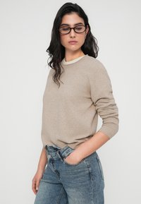 Μπεζ πουλόβερ με στρογγυλή λαιμόκοψη, μακριά μανίκια και ριμπ μανσέτες, συνδυασμένο με ανοιχτόχρωμο μπλε τζιν. Λεπτή υφή, άνετη εφαρμογή.