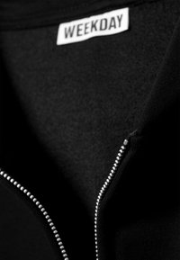 Giacca nera con cerniera a tutto capo, caratterizzata da una texture morbida, cerniera argentata e un'etichetta con la scritta "WEEKDAY" sul colletto.