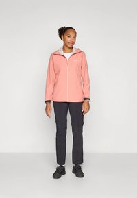 Lyserød, lynlåstrøje med høj krave, lange ærmer og sorte detaljer. Kombineret med mørke bukser og sorte sko.