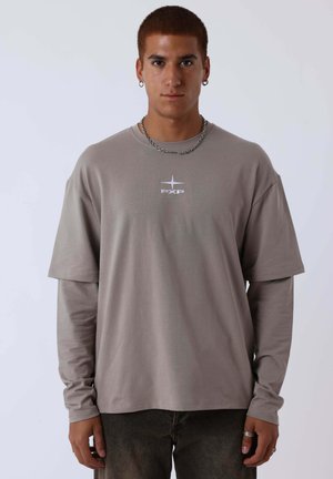 Jeune homme aux cheveux courts et bruns portant des chemises beige superposées à manches longues et courtes avec le logo "PXP", une chaîne argentée et des boucles d'oreilles anneaux.