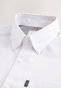 White button-down shirt collar with visible buttons, patterned inner collar lining, and a small navy tag with a white logo on the pocket.