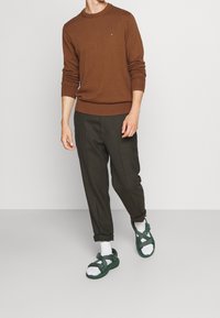 Homme portant un pull marron à manches longues, un pantalon foncé retroussé, des chaussettes blanches et des sandales vertes ouvertes, debout devant un fond uni.