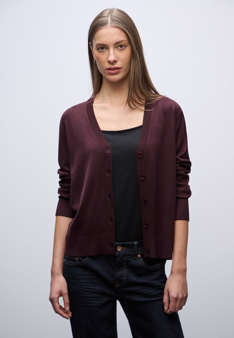 Cardigan a maglia borgogna con scollo a V, chiusura a bottoni e polsini a coste, indossato sopra una maglietta nera e jeans scuri. Design semplice e senza decorazioni.