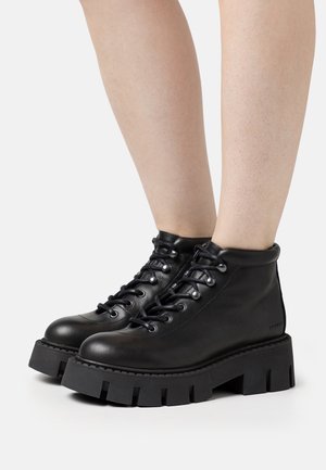 Sorte chunky platform støvler i læder med snørebånd foran, båret på bare ben mod hvid baggrund.