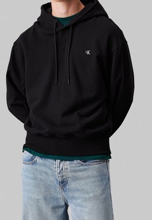 Mężczyzna w czarnej bluzie z kapturem Calvin Klein ze ściągaczami, na zielonej koszuli i jasnoniebieskich dżinsach na szarym tle.