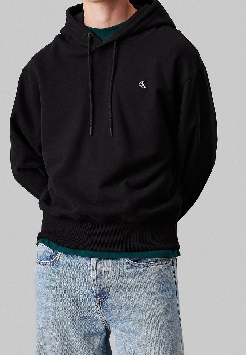 Homme portant un sweat à capuche noir Calvin Klein avec cordons sur une chemise verte et un jean bleu clair sur fond gris.