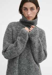 Siv sviterno z turtleneck-om z teksturiranim, puhastim zaključkom. Ima dolge rokave in ohlapen kroj, idealen za plastenje.