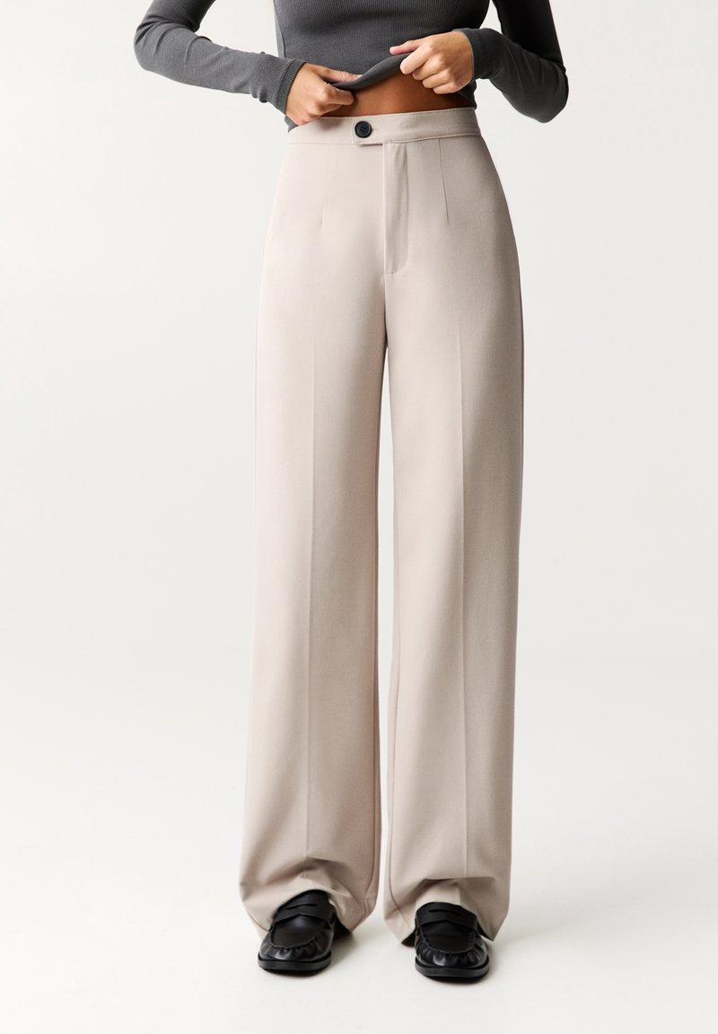 Personne portant un pantalon beige taille haute à jambes larges avec un bouton et des mocassins noirs, soulevant légèrement un haut à manches longues foncé pour montrer la taille.