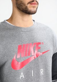 Harmaa huppari, jossa on suuri punainen Nike-logo, jossa on viiva ja sana "AIR" valkoisena. Pehmeä kangas, pyöreä pääntie ja pitkät hihat.