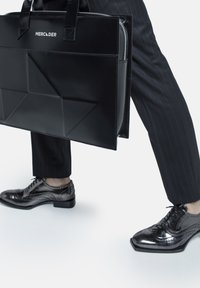 Black structured tote bag with diagonal folds, zip closure, and dual handles. Accompanied by polished black lace-up shoes with brogue detailing.