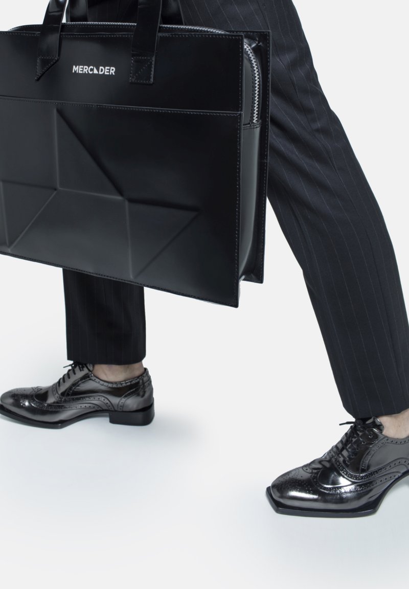 Black structured tote bag with diagonal folds, zip closure, and dual handles. Accompanied by polished black lace-up shoes with brogue detailing.