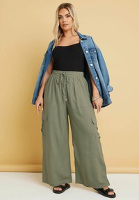 Olijfgroene wijde cargobroek met een elastische tailleband, gecombineerd met een zwarte tanktop en een spijkerjack. Zwarte sandalen met studs.