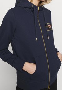 Námořnická modrá mikina na zip vyrobená z měkké tkaniny, s zlatým zipem, předními kapsami a vyšitým logem na hrudi.