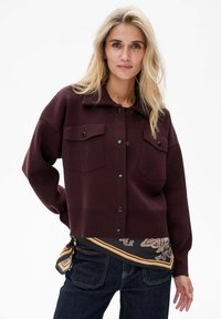 Kobieta o długich blond włosach, ubrana w ciemnoczerwony, zapinany na guziki żakiet, wzorzysty szalik przewiązany w pasie oraz ciemnoniebieskie dżinsy, stojąca na białym tle.