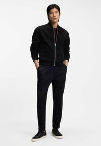 Black suede bomber jacket with a zip front, ribbed cuffs, and hem. Paired with tailored black trousers and black sneakers.
