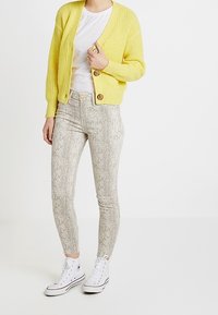 Cárdigan amarillo de punto con escote en V y cierre de botones. Camiseta blanca, pantalones ajustados con estampado de serpiente y zapatillas blancas de caña alta.