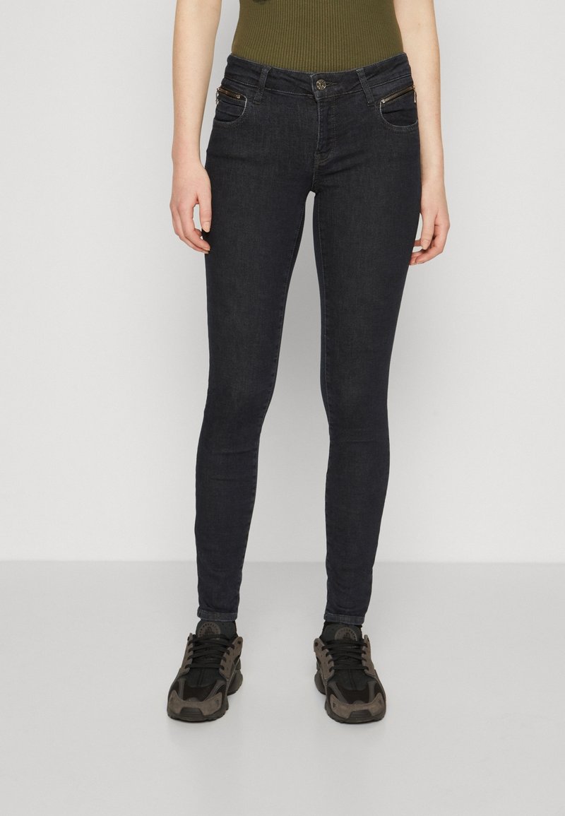 Jeans ajustados de denim oscuro con una textura suave, con bolsillos frontales con cremallera y una cintura de altura media, combinados con zapatillas negras.