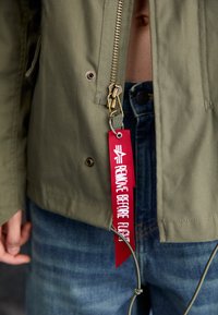 Olíva zöld dzseki cipzárral, amelyen egy piros szövetcímke található, rajta fehér betűkkel az áll, hogy "REMOVE BEFORE FLIGHT". Látható farmernadrág alatta.