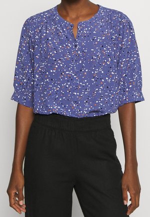 Nainen, joka on pukeutunut violettiin napilliseen puseroon, jossa on valkoisia, mustia ja oransseja pilkkuja, yhdistettynä korkeavyötäröisiin mustiin housuihin.