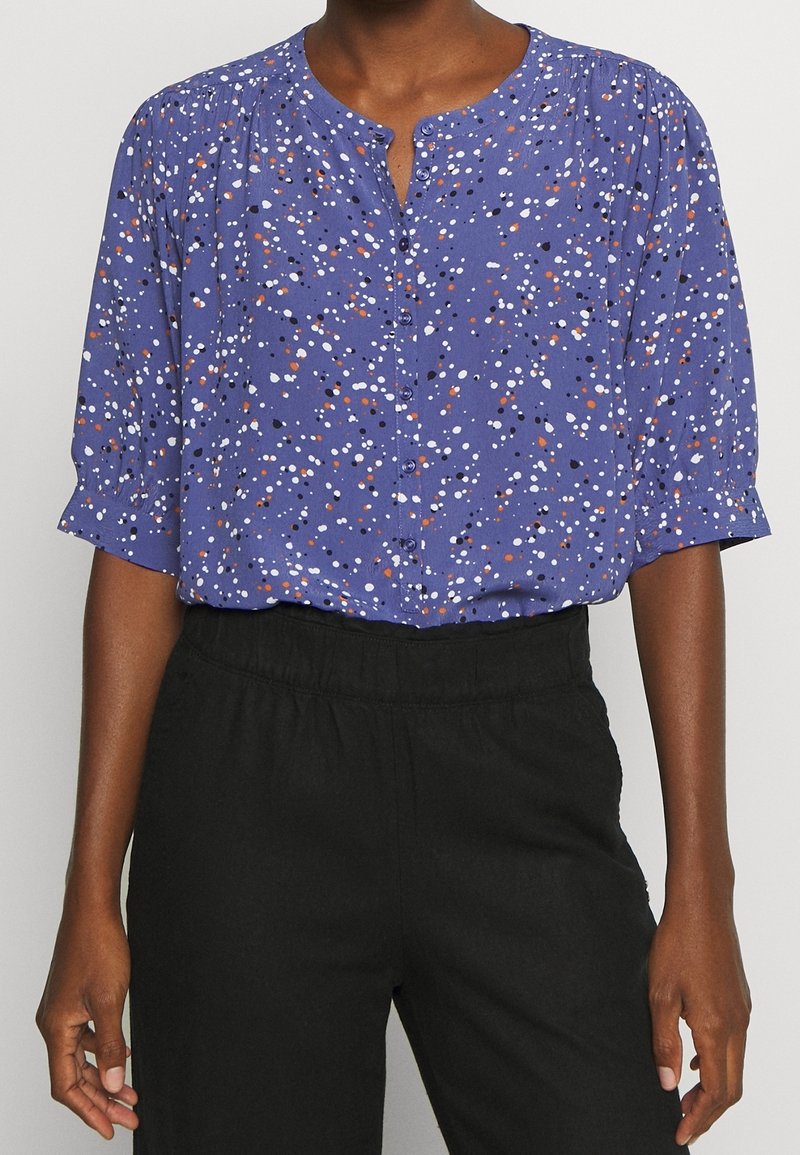 Femme portant un chemisier violet à boutons avec des taches blanches, noires et orange, associé à un pantalon noir taille haute.