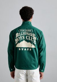 Grønn bomullsskjorte med et stort hvitt grafisk trykk på baksiden som har teksten "Billionaire Boys Club STAFF" og et fjelldesign.