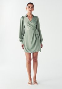 Zelené šaty na zaväzovanie s dlhými nafúknutými rukávmi, golierom a bočným viazaním; vyrobené z hladkej látky s jemným leskom. Biele sandále dokonale dopĺňajú vzhľad.