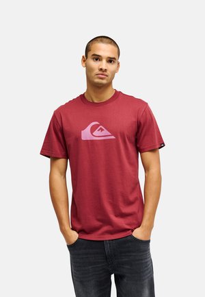 Junger Mann mit kurzen Haaren, der ein rotes T-Shirt mit einem pinken Wellen- und Berglogo trägt, die Hände in den Taschen, stehend vor einem schlichten Hintergrund.