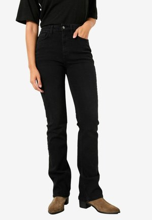 Schwarze hochtaillierte Jeans aus Denim mit einer figurbetonten Silhouette, Fronttaschen und einem leichten Bootcut. Kombiniert mit braunen Wildleder-Stiefeletten.
