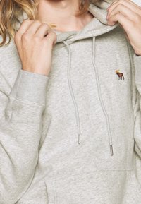 Grijze hoodie van zacht materiaal, met een buidelzak, een capuchon met koord en een klein geborduurd elandlogo op de linkerborst.