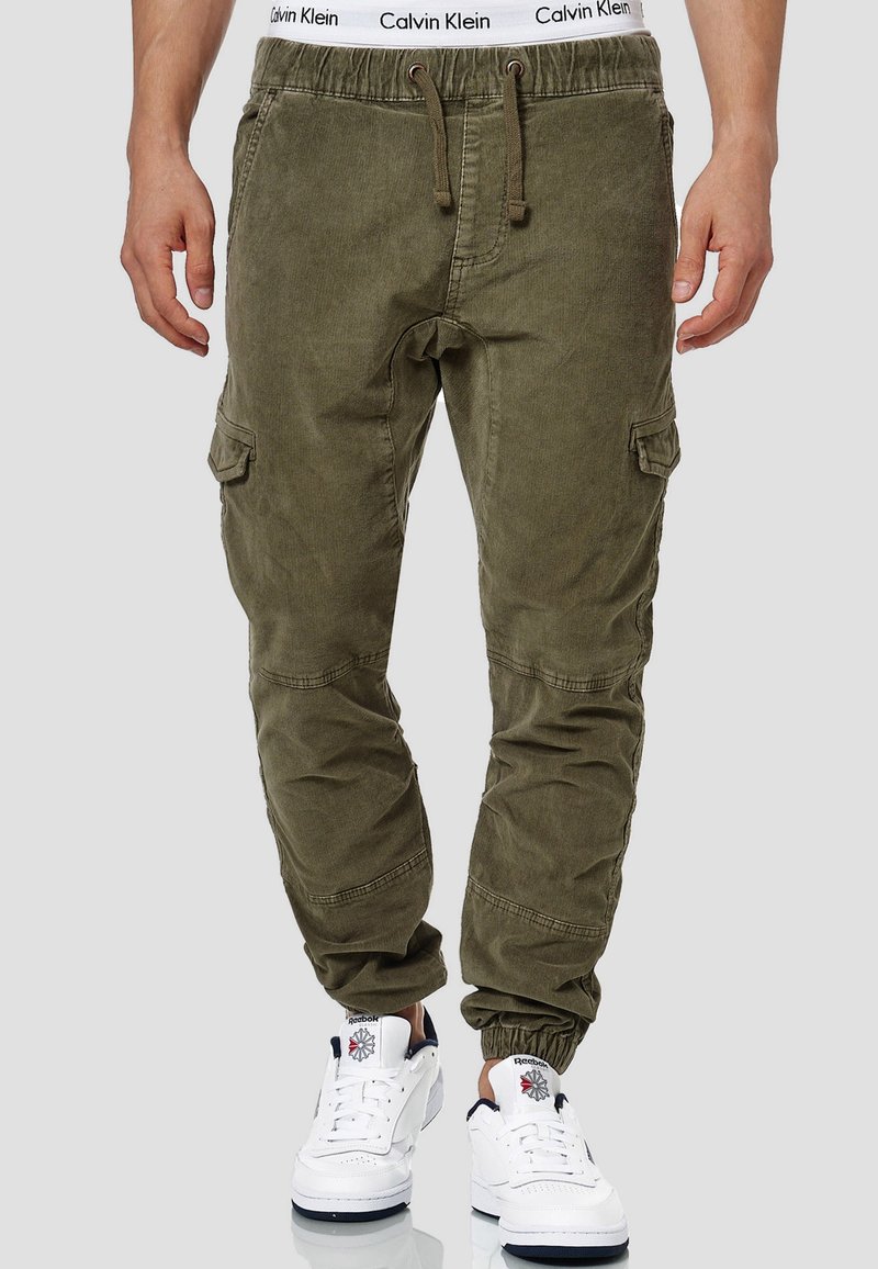 Homme portant un pantalon cargo jogger vert olive avec une taille à cordon, des poignets élastiques, une ceinture blanche Calvin Klein et des baskets blanches Reebok.