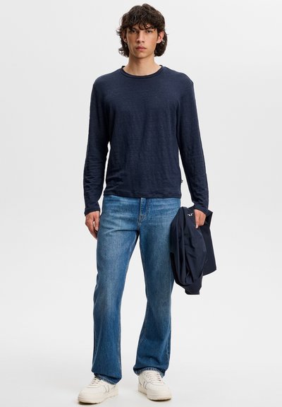Jeune homme aux cheveux bouclés portant une chemise à manches longues bleu marine, un jean bleu, des baskets blanches, tenant une veste foncée dans sa main gauche.