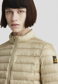 Giacca puffer beige con design a trapunta, colletto alto e cuciture verticali. Presenta una chiusura con zip e un patch logo sulla manica sinistra.