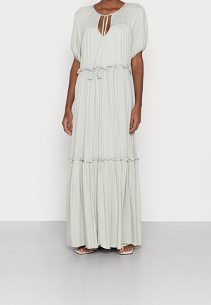Vrouw die een lichtgrijze maxi-jurk draagt met pofmouwen, een sleutelgat-halslijn met strikjes en gelaagde ruches, gecombineerd met witte sandalen met open teen.