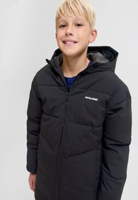 Melna polsterēta jaka ar rāvējslēdzēja aizdari un kapuci. Uz krūtīm ir redzams logo, un jaka ir ar šūtu rakstu, izgatavota no viegla sintētiska auduma.