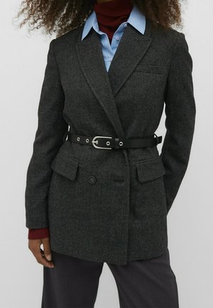 Personne portant un blazer croisé gris foncé avec une ceinture noire sur une chemise bleue et un col roulé rouge, debout devant un fond uni.