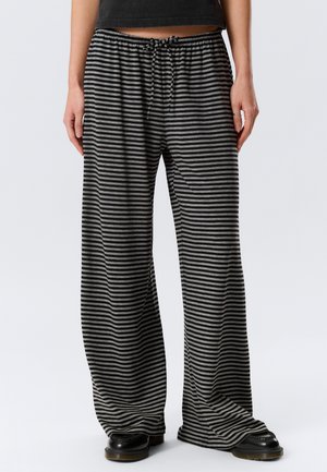 Osoba ubrana w szerokie spodnie z czarno-szarymi poziomymi paskami, z elastycznym pasem i czarnymi butami, stojąca na tle jednolitego tła.