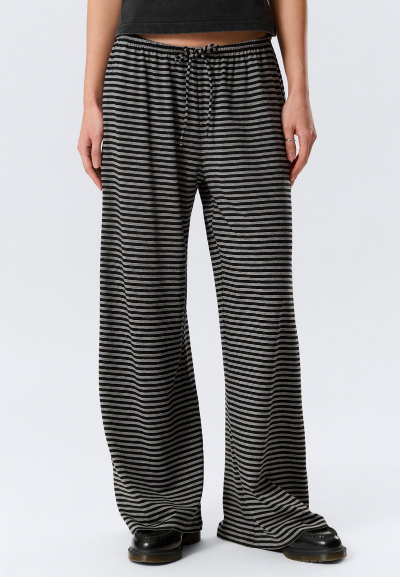 Άτομο που φορά μαύρο και γκρι παντελόνι με οριζόντιες ρίγες, φαρδύ και με λάστιχο στη μέση, και μαύρα παπούτσια, στέκεται μπροστά σε απλό φόντο.