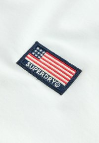 Bijela tkanina s pravokutnom izvezenom oznakom na kojoj je američka zastava i tekst "SUPERDRY®" u sivoj boji na tamno plavoj pozadini.