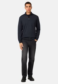 Moška mornarsko modra zadrgo zapeta jopica iz pletenega materiala z visokim ovratnikom in dolgimi rokavi, v kombinaciji s temnimi jeans in črnimi supergami.